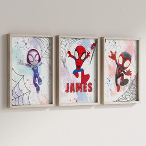 May include: Three framed prints featuring cartoon superheroes in a colorful, abstract style. The prints feature a female superhero in purple and blue, a male superhero in red and blue, and a male superhero in black and red. The middle print includes the name "JAMES" in red letters.