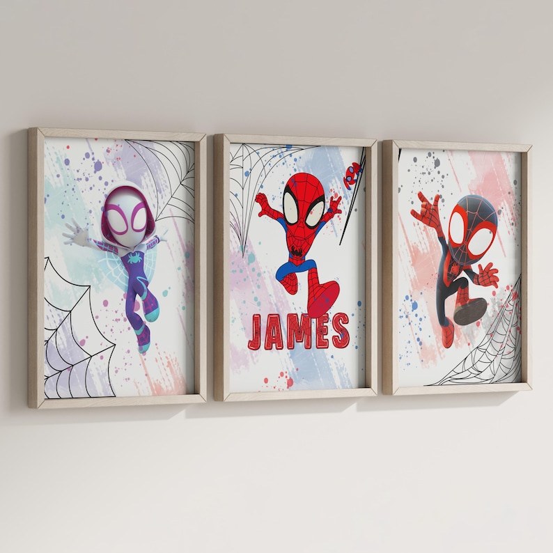 May include: Three framed prints featuring cartoon superheroes in a colorful, abstract style. The prints feature a female superhero in purple and blue, a male superhero in red and blue, and a male superhero in black and red. The middle print includes the name "JAMES" in red letters.