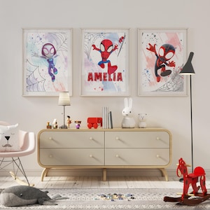 May include: Three framed prints featuring a cartoon superhero in a red and blue suit with a spider design. The prints are hung on a white wall above a dresser with four drawers. The middle print has the name "Amelia" written on it.