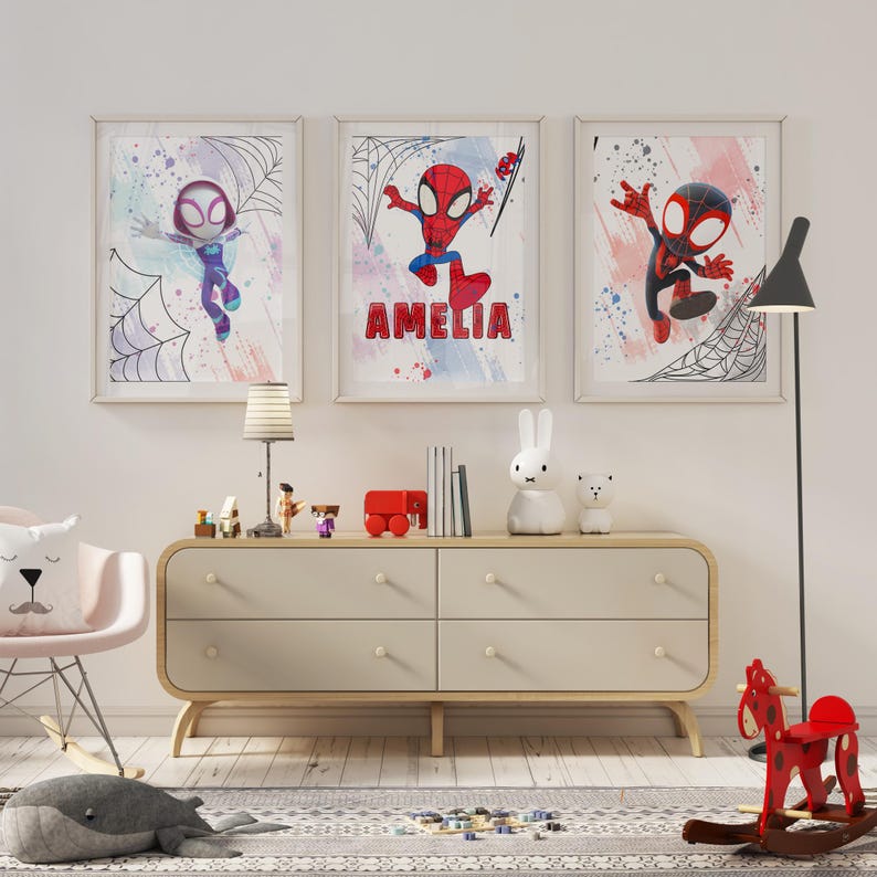 May include: Three framed prints featuring a cartoon superhero in a red and blue suit with a spider design. The prints are hung on a white wall above a dresser with four drawers. The middle print has the name "Amelia" written on it.