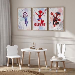 May include: Three framed prints of cartoon superheroes with a white background and colorful watercolor splatters. The prints feature a red and blue Spiderman, a black and red Spiderman, and a purple and blue Spiderman. The middle print has the name "Oliver" written in red letters.
