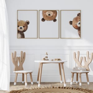 May include: Three framed prints of brown bear faces, two looking to the right and one upside down, hanging on a white wall above a white table and two wooden chairs with antlers.