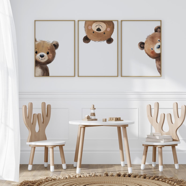 May include: Three framed prints of brown bear faces, two looking to the right and one upside down, hanging on a white wall above a white table and two wooden chairs with antlers.