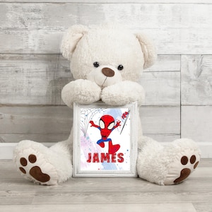 May include: A white teddy bear holding a framed print of a cartoon Spiderman in a red and blue suit. The print has a blue, pink, and white watercolor background with the name "JAMES" written in white.