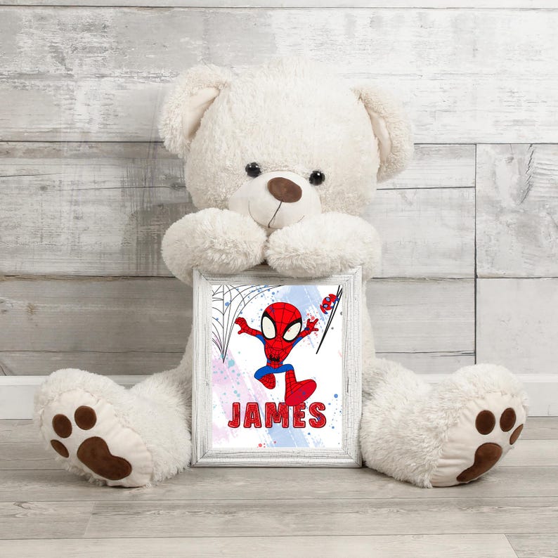 May include: A white teddy bear holding a framed print of a cartoon Spiderman in a red and blue suit. The print has a blue, pink, and white watercolor background with the name "JAMES" written in white.