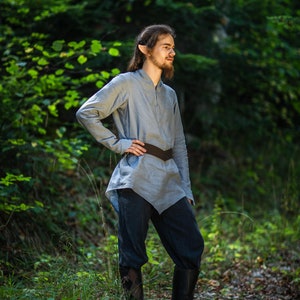 May include: A person wearing a light blue tunic with a brown belt and black pants, standing in a forest setting.