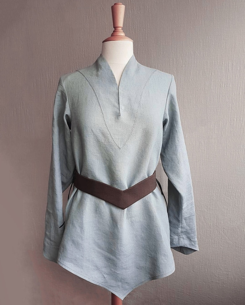 May include: A light blue linen tunic with a V-neck and long sleeves. The tunic has a brown leather belt with a pointed front.