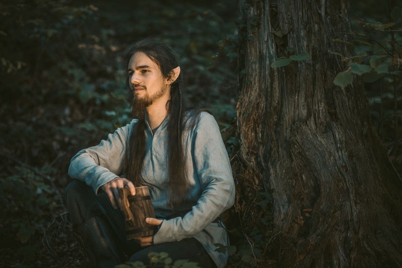 May include: A man with long, dark hair and pointed ears sits in a forest, holding a wooden mug. He is wearing a light blue shirt and dark pants. The background is a dark green forest with a large tree trunk.