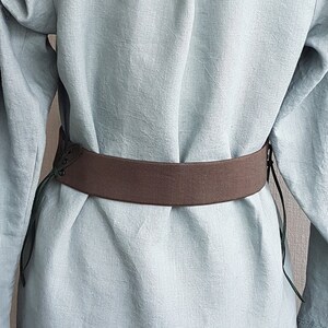 May include: A brown fabric belt with green laces. The belt is tied around a light blue linen shirt.