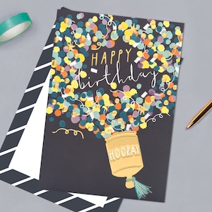 May include: A black card with a gold and colourful confetti design. The confetti is coming out of a gold container with the word "HOORAY" on it. The text "Happy birthday" is written in white on the card.