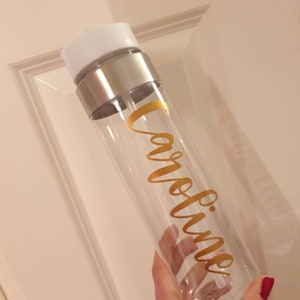 Puede incluir: Una botella de agua de pl&aacute;stico transparente con una tapa plateada y una pegatina de vinilo dorada que dice "Caroline".