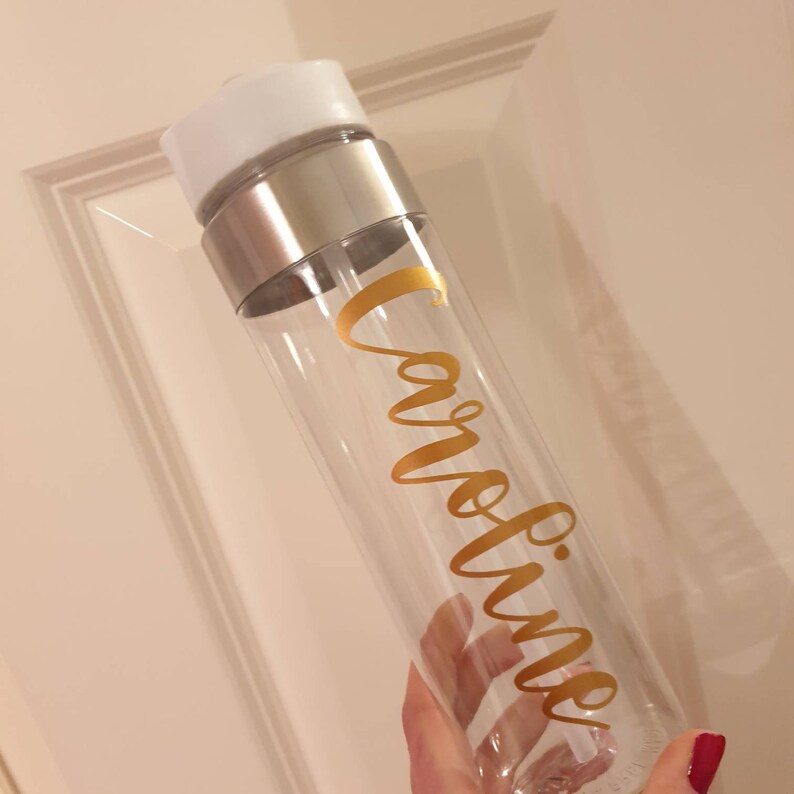 Puede incluir: Una botella de agua de pl&aacute;stico transparente con una tapa plateada y una pegatina de vinilo dorada que dice "Caroline".