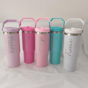 May include: Five stainless steel tumblers with carrying handles and lids, each featuring a unique name engraved vertically: Maddie, Naomi, Anya, Isaac, and Kate. The tumblers are coloured in lavender, hot pink, light pink, turquoise, and white.