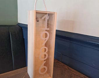 Puede incluir: Una caja de vino de madera alta y rectangular con un panel frontal de acrílico transparente. El panel presenta el texto vertical 1986 y la frase On your 40th. La caja incluye un asa de cuerda de color claro unida a la parte superior.
