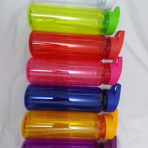 Puede incluir: Conjunto de siete botellas de agua de pl&aacute;stico transparente con tapas de diferentes colores. Las tapas tienen un dise&ntilde;o de tapa abatible. Las botellas est&aacute;n dispuestas en una pila vertical de arriba a abajo: transparente, amarilla, roja, rosa, azul, naranja y morada.