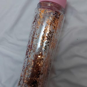 Puede incluir: Una botella de agua de pl&aacute;stico transparente con una tapa rosa y un dise&ntilde;o de purpurina dorada.