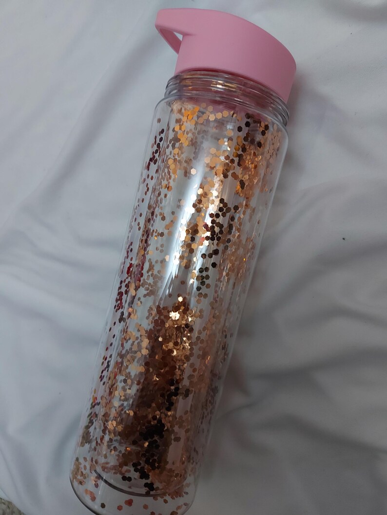 Puede incluir: Una botella de agua de pl&aacute;stico transparente con una tapa rosa y un dise&ntilde;o de purpurina dorada.