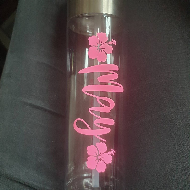 Puede incluir: Una botella de agua de pl&aacute;stico transparente con tapa plateada y una pajita rosa. La botella tiene un dise&ntilde;o de flores de hibisco rosas y el nombre "Molly" escrito en cursiva rosa.