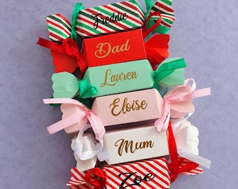 Puede incluir: Una pila de seis cajas de regalo con forma de caramelo en varios colores, incluyendo rojo, verde, rosa y blanco, cada una atada con cintas a juego. Las cajas están personalizadas con los nombres Freddie, Dad, Lauren, Eloise, Mum y Zoe en cursiva.