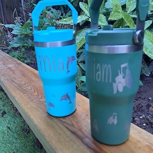 May include: Two personalised insulated tumblers with handles. One is teal with the name "mia" and dolphin graphics. The other is green with the name "william" and a tractor graphic. Both tumblers are on a wooden surface.
