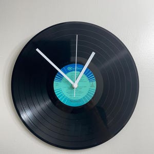Pode incluir: Um relógio circular feito de um disco de vinil preto, com ponteiros brancos. O rótulo central é azul e verde-azulado, com a palavra "Camden" e outro texto. Os ponteiros do relógio indicam 10:08. A palavra "MONO" está impressa no rótulo.