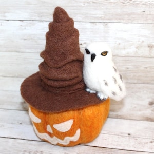 May include: A felted Halloween decoration featuring an orange pumpkin with a carved face, a brown witch's hat, and a white owl with yellow eyes. The owl is perched on the hat. The background is a light wood grain.