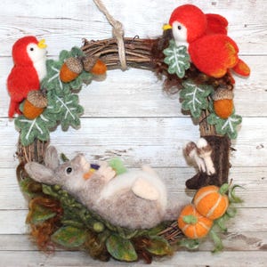 May include: A decorative wreath made of woven twigs featuring two needle-felted red birds, a grey rabbit lying on its back, green felt leaves, brown acorns, and two small orange pumpkins. The wreath is suspended by a light-colored rope loop.