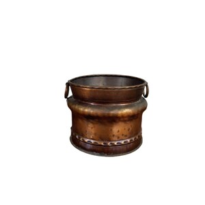 May include: A copper-coloured metal planter with a round, open top and two small handles. The planter has a wide, cylindrical body with a decorative band around the middle. The metal has a slightly aged appearance.