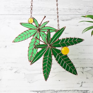 May include: A stained glass suncatcher featuring two green leaves with yellow glass beads. The leaves are connected by a copper chain.
