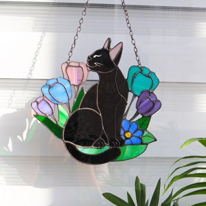 May include: A stained glass suncatcher featuring a black cat sitting amongst colorful tulips and a blue forget-me-not flower. The cat is looking to the right and the suncatcher is hanging from a chain.