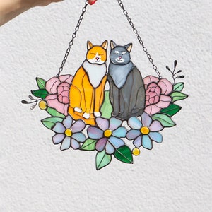 Op de afbeelding: Een gebrandschilderd raam dat aan een ketting hangt, met twee katten die tussen roze en paarse bloemen zitten. De oranje kat zit links en de grijze kat rechts.