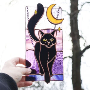 May include: A stained glass window hanging features a black cat with yellow eyes standing in front of a crescent moon. The background is purple and pink.