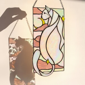 May include: A stained glass window hanging on a chain, depicting a white cat with a crescent moon and stars in a pink and green landscape.