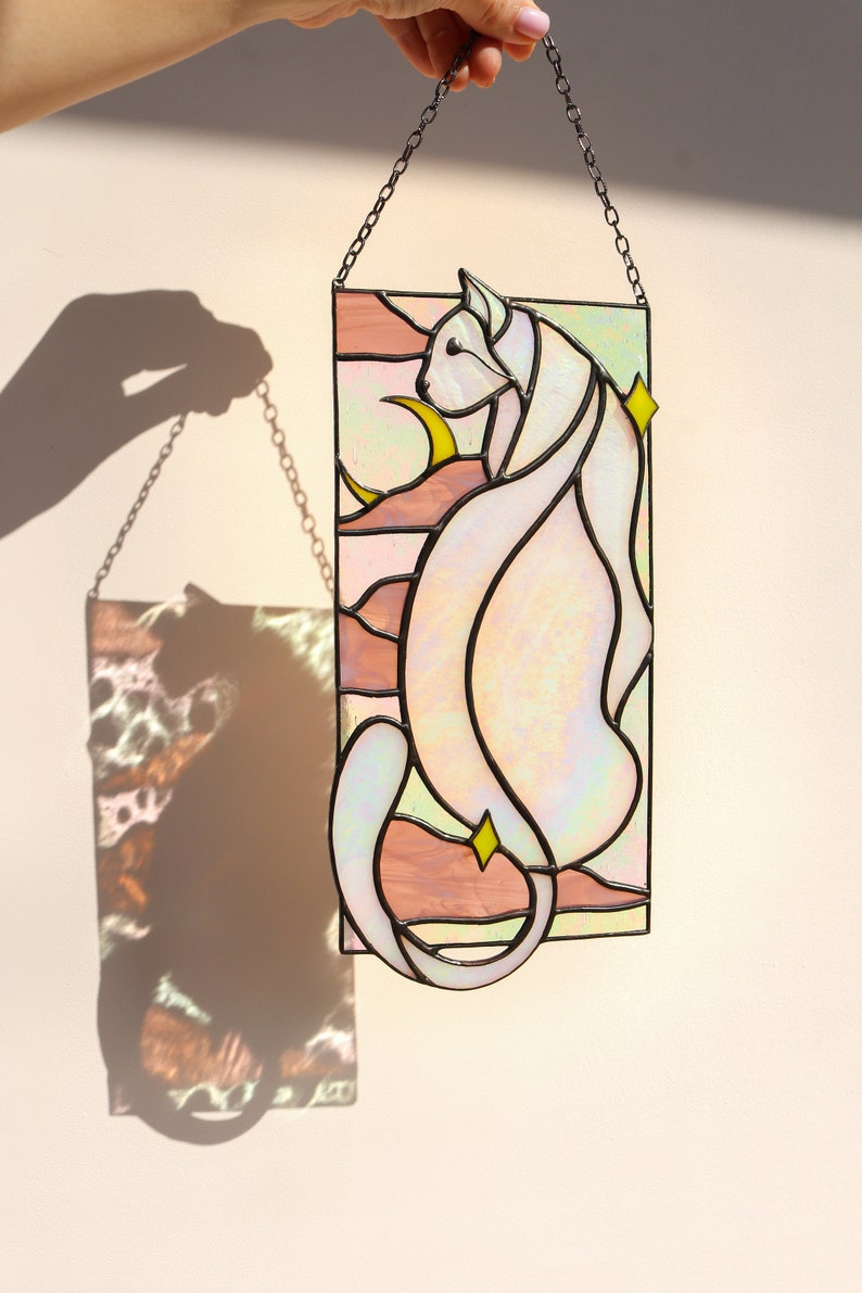 May include: A stained glass window hanging on a chain, depicting a white cat with a crescent moon and stars in a pink and green landscape.