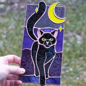 May include: A stained glass window panel depicting a black cat with yellow eyes standing in front of a crescent moon and stars on a purple background.