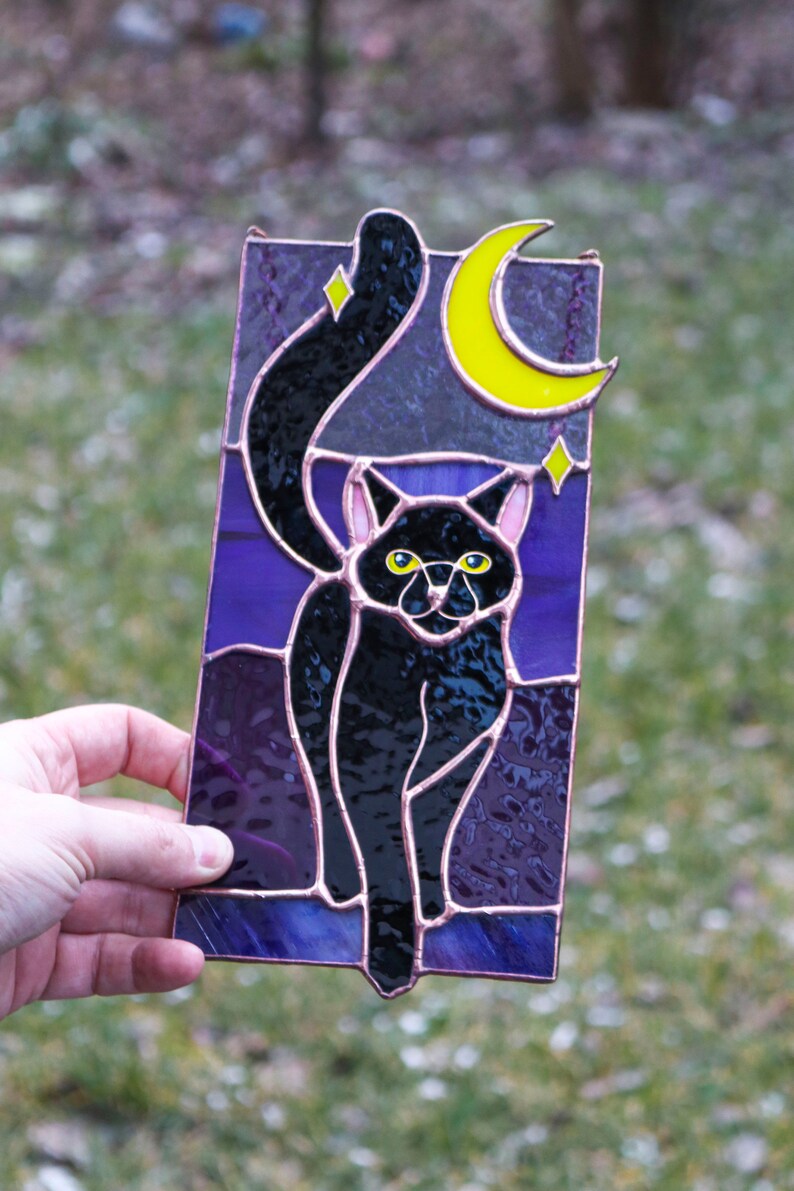 May include: A stained glass window panel depicting a black cat with yellow eyes standing in front of a crescent moon and stars on a purple background.