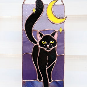 May include: A stained glass window hanging with a black cat silhouette with yellow eyes, a crescent moon, and stars against a purple background.