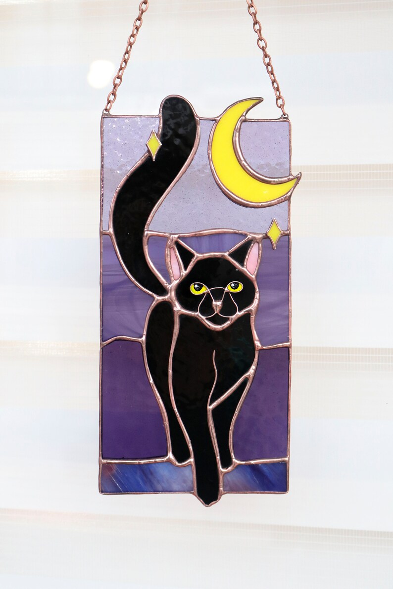 May include: A stained glass window hanging with a black cat silhouette with yellow eyes, a crescent moon, and stars against a purple background.
