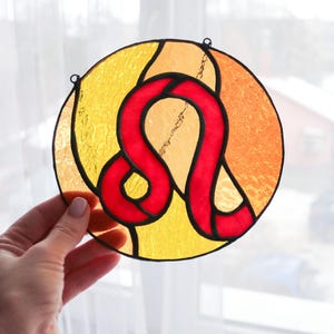 Könnte beinhalten: Ein rundes Buntglasstück mit dem Sternzeichen Löwe in Rot auf gelb-orangem Hintergrund. Das Kunstwerk hat eine schwarze Umrandung und einen kleinen Metallring zum Aufhängen.