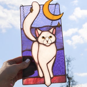 May include: A stained glass window hanging with a white cat with yellow eyes, a crescent moon, and stars on a purple background.