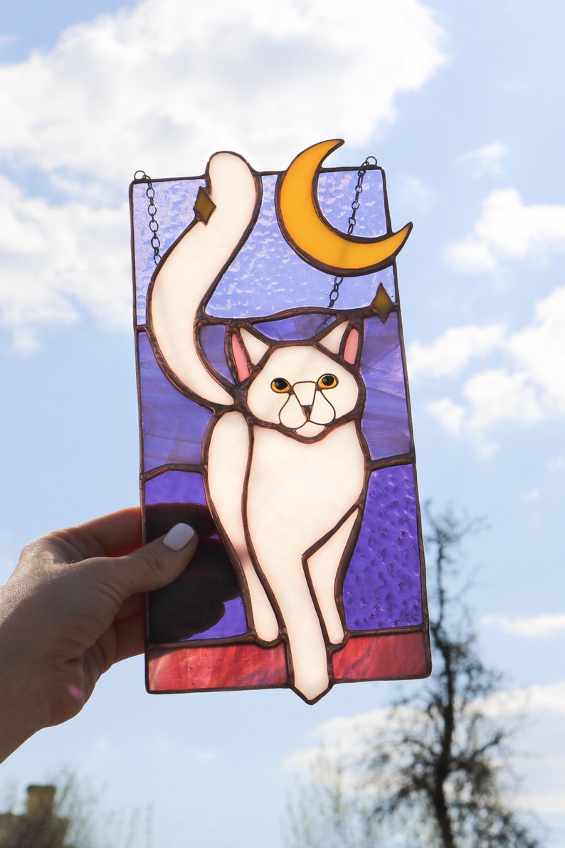 May include: A stained glass window hanging with a white cat with yellow eyes, a crescent moon, and stars on a purple background.