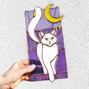 May include: A stained glass window hanging with a white cat with yellow eyes, a yellow crescent moon, and stars on a purple background.