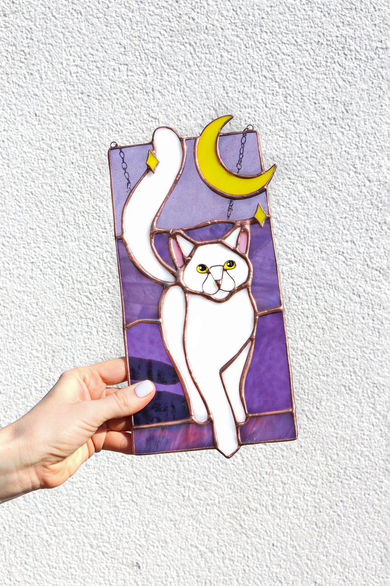 May include: A stained glass window hanging with a white cat with yellow eyes, a yellow crescent moon, and stars on a purple background.
