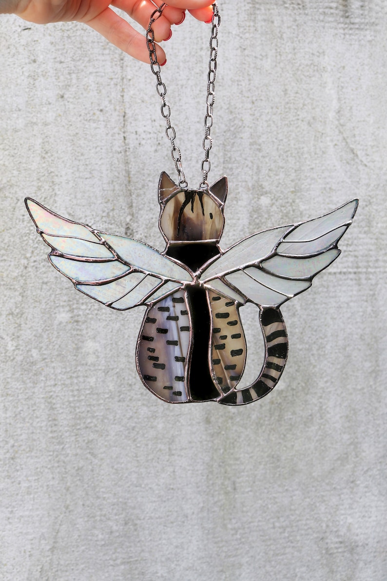 May include: A stained glass cat with wings hanging from a silver chain. The cat is black and brown with white stripes. The wings are iridescent.
