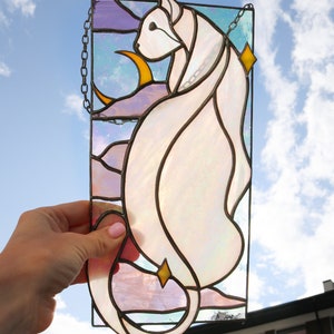 May include: A stained glass window hanging with a chain, depicting a white cat with a crescent moon and stars in the background. The cat is outlined in black and the moon and stars are yellow.