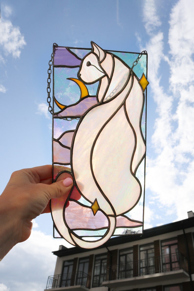 May include: A stained glass window hanging with a chain, depicting a white cat with a crescent moon and stars in the background. The cat is outlined in black and the moon and stars are yellow.