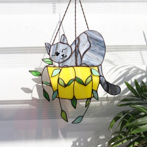 May include: A stained glass hanging decoration featuring a gray cat sitting in a yellow planter with green leaves. The cat is looking to the right and has a cute expression on its face.