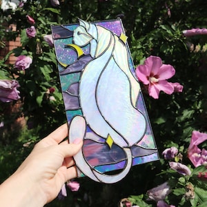 May include: A stained glass window panel featuring a white cat with iridescent glass, sitting in a crescent moon with stars. The background is a purple, blue, and green gradient.