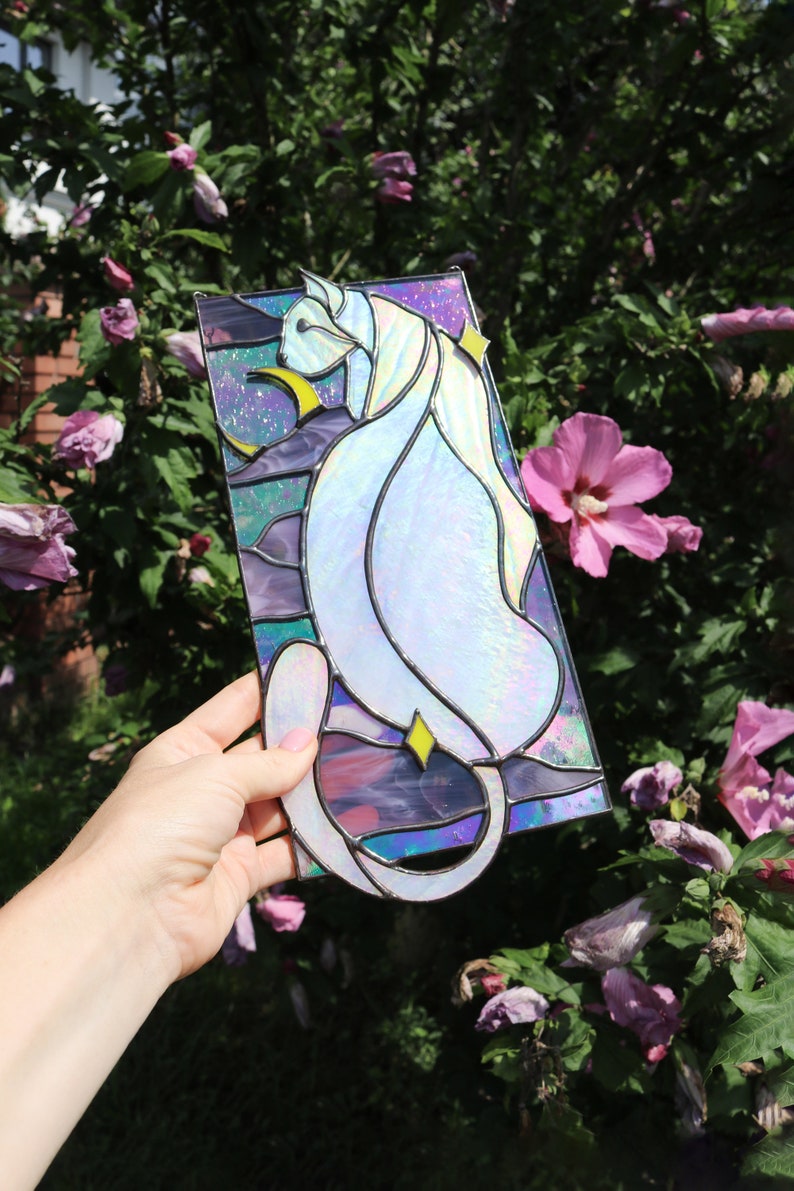 May include: A stained glass window panel featuring a white cat with iridescent glass, sitting in a crescent moon with stars. The background is a purple, blue, and green gradient.