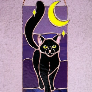 May include: A stained glass window hanging with a black cat silhouette walking in front of a purple night sky with a yellow crescent moon and stars.